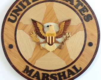 Op de afbeelding: Een houten plaquette met een diameter van 35,5 cm met een vijfpuntige ster met een adelaar- en schildontwerp. De plaquette is gemaakt van verschillende soorten hout en heeft een donkerbruine rand. De tekst "UNITED STATES MARSHAL" staat in donkerbruine letters rond de ster. Het schild heeft de tekst "1789" erop.