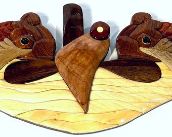 May include: Wooden wall art featuring two fish flanking a central emblem. The fish have dark red scales and light brown fins. The piece measures 21 inches long and 6.5 inches high. Officially Licensed by the U.S. Navy.