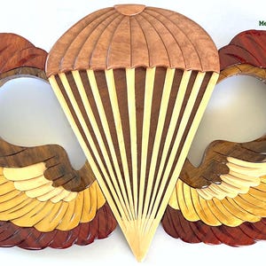 May include: Wooden wall art featuring a parachute and wings design. The artwork measures 18 inches by 10 inches. The parachute is light brown with cream-colored stripes, and the wings are a combination of brown, yellow, and red wood.