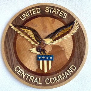 May include: A wooden plaque with a circular design featuring a bald eagle with outstretched wings holding a shield with the American flag. The plaque is inscribed with the words "United States Central Command".