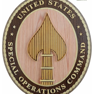 May include: A round wooden plaque with a brown border and a light brown center. The plaque features the United States Special Operations Command logo, which includes a gold spearhead with a column base and the words "United States Special Operations Command". The plaque measures 16"L x 14"W.