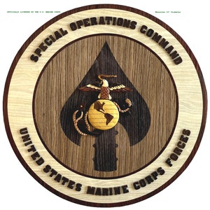 Puede incluir: Una placa de madera con un acabado de grano de madera marrón y negro. La placa presenta el logotipo del Comando de Operaciones Especiales del Cuerpo de Marines de los Estados Unidos, que incluye un globo terráqueo, un águila y un ancla. El texto "SPECIAL OPERATIONS COMMAND" está escrito en un patrón circular alrededor del logotipo. El texto "UNITED STATES MARINE CORPS FORCES" está escrito en un patrón circular alrededor del logotipo. La placa mide 35,56 cm de diámetro.