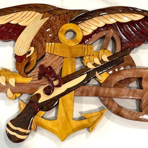 May include: Wooden wall art featuring a detailed eagle with outstretched wings, an anchor, a trident, and a rifle. The artwork uses various wood tones, including brown, red, and yellow, creating a layered effect. The eagle's wings are a mix of red and cream.