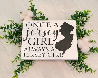 May include: A white wooden sign featuring the text "ONCE A JERSEY GIRL ALWAYS A JERSEY GIRL" in black script, with a black silhouette of New Jersey. The sign is decorated with green foliage and cotton.