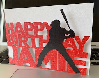 May include: A handmade birthday card with a baseball theme. The card features the words "HAPPY BIRTHDAY JAMES" cut out in red, with a black silhouette of a baseball player swinging a bat.
