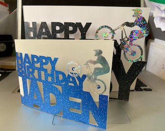 May include: A handmade birthday card with a blue and white design. The card features the words "HAPPY BIRTHDAY JADEN" cut out in a blue glittery font. A holographic image of a person riding a bicycle is also included.