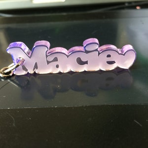 May include: A keychain with the name "Macie" in a purple and white bubble letter font. The keychain is made of a translucent material and has a silver chain attached. The name is outlined in a darker purple.