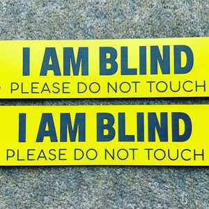 May include: Two rectangular yellow signs with rounded corners. The signs have the text "I AM BLIND PLEASE DO NOT TOUCH" in dark blue. Paw prints are on each sign. Each sign has two holes for mounting.