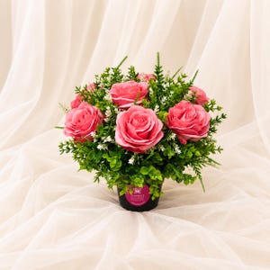 May include: A floral arrangement featuring multiple vibrant pink roses nestled among dense green foliage and small white filler flowers. The flowers are contained in a black pot with a circular pink label displaying the text Sianee and butterfly icon.