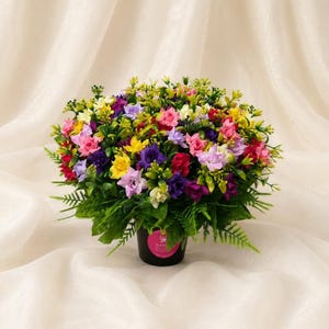 May include: A dense, rounded arrangement of artificial flowers in a black pot. The bouquet features a vibrant mix of small blossoms in shades of pink, yellow, purple, red, and white, interspersed with green foliage and fern-like leaves.