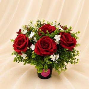 May include: A floral arrangement featuring deep red roses and small white blossoms nestled among dense green foliage. The flowers are contained in a black cylindrical pot with a pink circular label that displays the text Sianee's.