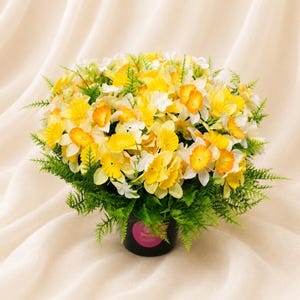May include: A dense bouquet of artificial yellow and white daffodils with green fern-like foliage arranged in a black cylindrical pot. The pot features a circular pink label with the text Sianee printed in white on the front of the container.