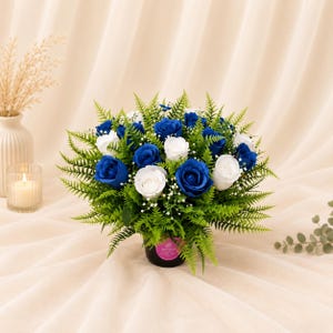 May include: A floral arrangement featuring vibrant blue and white roses interspersed with lush green fern fronds and small white filler flowers. The bouquet is contained in a dark cylindrical base with a small pink label, creating a dense, rounded display.