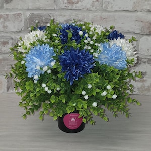 May include: A decorative floral arrangement with blue, white, and light blue artificial flowers. The flowers are set in a bed of green foliage with small white accents, in a black pot. The background is a brick wall.