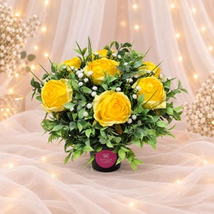 May include: A bouquet of bright yellow roses arranged with small white flowers and green foliage in a black pot. The pot features a circular pink label with a butterfly icon and the text Sianee's New Flowers New Creations printed in white.