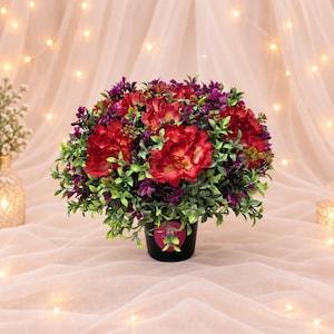 May include: A round artificial floral arrangement featuring vibrant red carnations mixed with deep purple and green foliage. The flowers are contained in a simple black cylindrical pot with a small circular pink label on the front side.