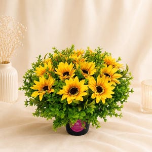 May include: A round artificial floral arrangement featuring bright yellow sunflowers with dark brown centres, surrounded by dense green foliage. The flowers are contained in a small black pot with a circular pink label on the front side.