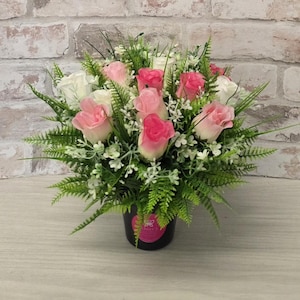 May include: A floral arrangement with pink and white roses, green fern-like foliage, and small white flowers. The flowers are in a black pot, creating a colourful display. The background is a brick wall.