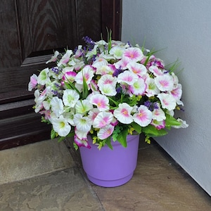 May include: A colourful display of artificial flowers in a lilac pot. The flowers are white, pink, and purple, with green leaves. The pot is round and sits on a stone surface, suitable for home decoration.