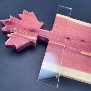 May include: A wooden cutting board shaped like a maple leaf with a clear acrylic insert. The board is made of cedar wood and has a natural wood grain.
