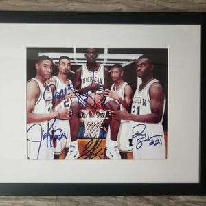 May include: Framed photograph of a basketball team in white and yellow jerseys, with signatures in blue ink. The team is posed with a basketball and net. The frame is black.