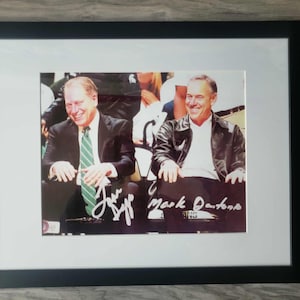 May include: Framed photograph featuring two smiling men, one in a suit with a green tie and the other in a leather jacket. The photo is signed with the names "Jeff" and "Mark Dantonio" in white ink.