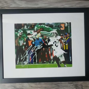 May include: Framed photograph of a football player in a green uniform leaping over a player in a white uniform. The image is signed in silver ink. The background shows other players and the number 3.