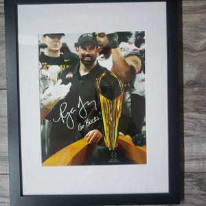 May include: Framed photograph featuring a smiling man holding a trophy, with a signature and the words "Go Bucks!". The trophy is gold and black. The photograph is in a black frame with a white mat.