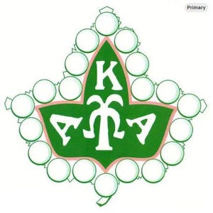 Puede incluir: Un emblema verde y blanco en forma de hoja, con un borde rosa. El emblema presenta las letras griegas K, M y A en blanco, junto con un diseño blanco estilizado. La hoja está rodeada de círculos blancos.