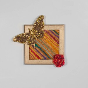 Peut inclure: Une œuvre d'art encadrée avec une peinture abstraite colorée, un papillon en paillettes dorées et une fleur rouge en crochet.