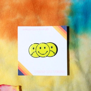 May include: A yellow enamel pin with three smiley faces, one sad, one happy, and one sad. The pin is on a white card with the text "PINS BY TESS" in pink.