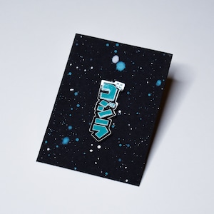 May include: A teal blue enamel pin with the Japanese word "Godzilla" written in white letters. The pin is on a black background with white speckles.