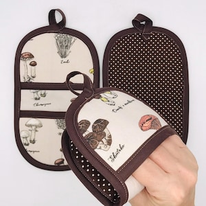 May include: Set of three brown and cream-colored oven mitts and pot holders. One mitt features a white background with mushroom illustrations and text. The other two have a brown polka dot and mushroom illustration design.