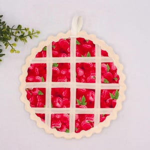 May include: A round fabric potholder featuring a vibrant red raspberry print with green leaves. The surface is overlaid with a cream-colored lattice grid pattern and finished with a scalloped edge and a small fabric hanging loop at the top.