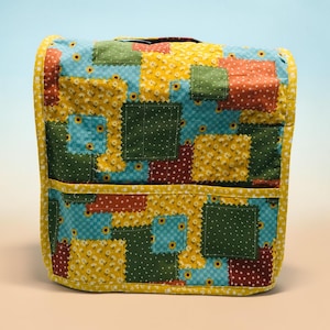 May include: A quilted fabric cover featuring a patchwork design of squares in yellow, green, teal, and dark red. The pattern includes floral prints, small dots, and checkered textures, finished with a solid yellow border and a horizontal pocket.