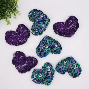 May include: Seven heart-shaped fabric ornaments in shades of purple, blue, and green. Some hearts feature a floral pattern, while others have a dotted design. The ornaments are arranged on a white surface, with a sprig of green foliage in the upper left corner.