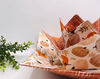 May include: A fabric bowl cosy featuring a cream-coloured exterior with a repeating pattern of orange and brown pumpkins and autumn leaves. The interior and edges are lined with a burnt orange fabric featuring a white grid pattern.