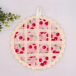 May include: A round fabric potholder featuring a pink background with a repeating cherry pattern and green leaves. The surface is overlaid with a cream-colored lattice grid design, finished with a scalloped edge and a small fabric hanging loop.