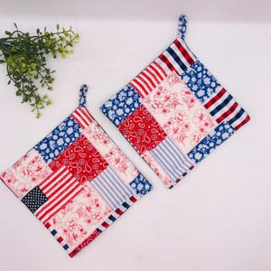 May include: Two square fabric pot holders featuring a patchwork design of red, white, and blue patterns. The fabric includes motifs of American flags, floral prints, paisley, and stripes. Each pot holder has a small fabric loop for hanging.