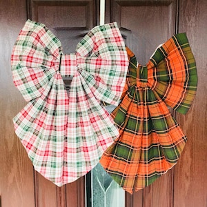 May include: Two large decorative bows made of fabric with plaid patterns. One bow features a white base with red and green intersecting lines. The second bow features an orange base with dark green and black intersecting plaid lines.