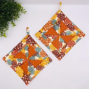May include: Two square quilted potholders featuring a pinwheel design in the center made of solid orange fabric with white dots. The surrounding fabric displays a floral pattern with yellow, maroon, and teal petals on a cream background.
