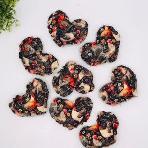 May include: Heart-shaped fabric coasters with a rooster and floral pattern. The coasters feature a dark background with colorful roosters and flowers. The coasters are made of fabric and are likely used to protect surfaces from heat or moisture.