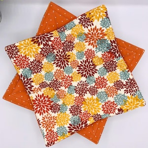 May include: Two square quilted pot holders. The top pot holder has a cream background with a floral pattern in shades of yellow, red, and blue. The bottom pot holder is solid orange with white polka dots.