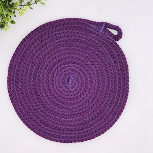 Könnte beinhalten: Ein runder, gewebter Untersetzer in Lila mit einer Schlaufenschlaufe. Der Untersetzer besteht aus dickem, geflochtenem Material und hat ein Spiraldesign. Die Farbe ist ein tiefes, sattes Lila. Der Untersetzer dient zum Schutz von Oberflächen vor heißen Gegenständen.