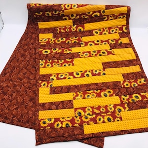 May include: A quilted table runner featuring a patchwork design. The runner has alternating sections of yellow and sunflower-patterned fabric, with a brown leaf-patterned border. The color palette includes shades of yellow, brown, and red.