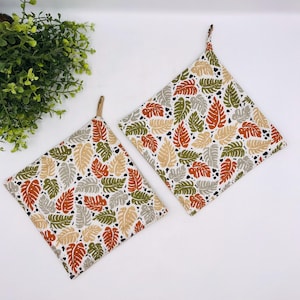 May include: Two square pot holders with a leaf pattern in shades of orange, green, tan, and gray on a white background. Each pot holder has a loop for hanging. The leaf design is repeated across the fabric.