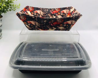 May include: A fabric bowl with a rooster design in black and red sits above a black rectangular food container with a clear lid. The bowl is open and angled, displaying the rooster and floral pattern.