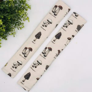 May include: Two beige fabric seat belt covers with a repeating pattern of various mushroom illustrations and text. The mushrooms include Red cap boletus, Champignon, Morel, and King bolete.