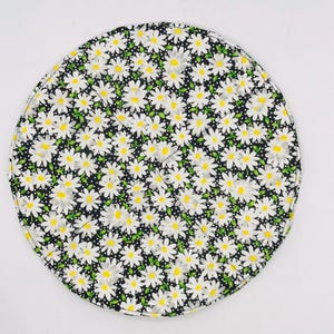 Op de afbeelding: Een rond, gewatteerd stoffen item met een zwarte achtergrond bedekt met een madeliefjespatroon. De madeliefjes hebben witte bloemblaadjes, gele centra en groene bladeren. Het item is waarschijnlijk een placemat of decoratief stuk.
