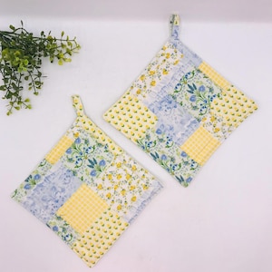 May include: Two square quilted potholders featuring a patchwork design of yellow and blue floral patterns, yellow gingham checks, and light blue damask prints. Each potholder includes a small fabric loop at the corner for convenient hanging.